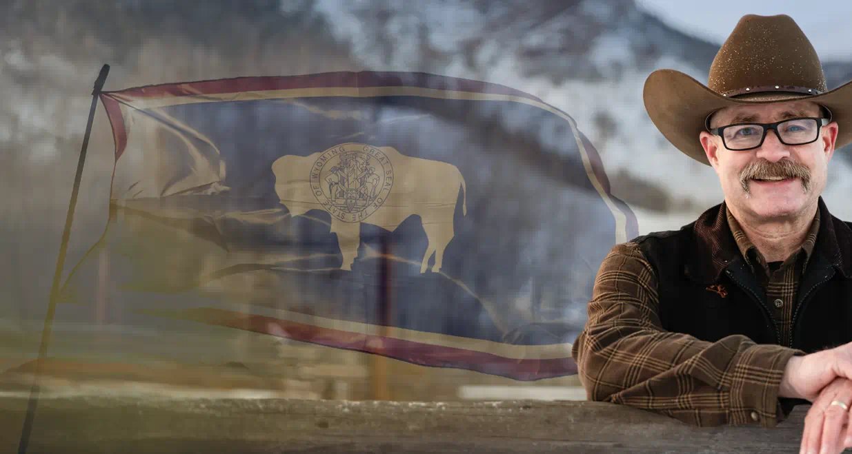 Daron L. Wyatt leaning on a wooden fence rail, with the Wyoming state flag and snow-covered hills behind him